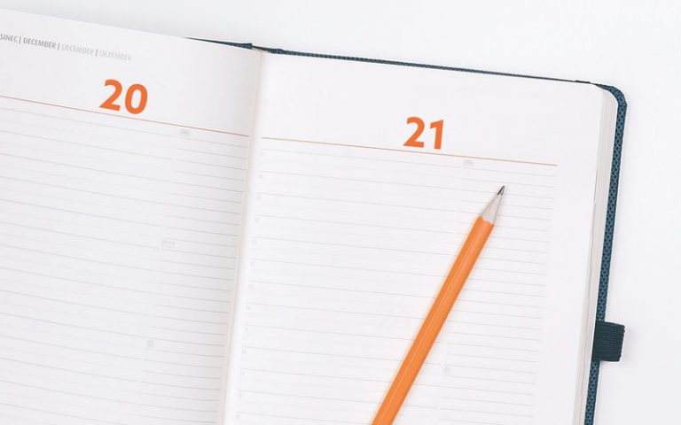 Aufgeschlagener Kalender mit orangenem Bleistift oben drauf.