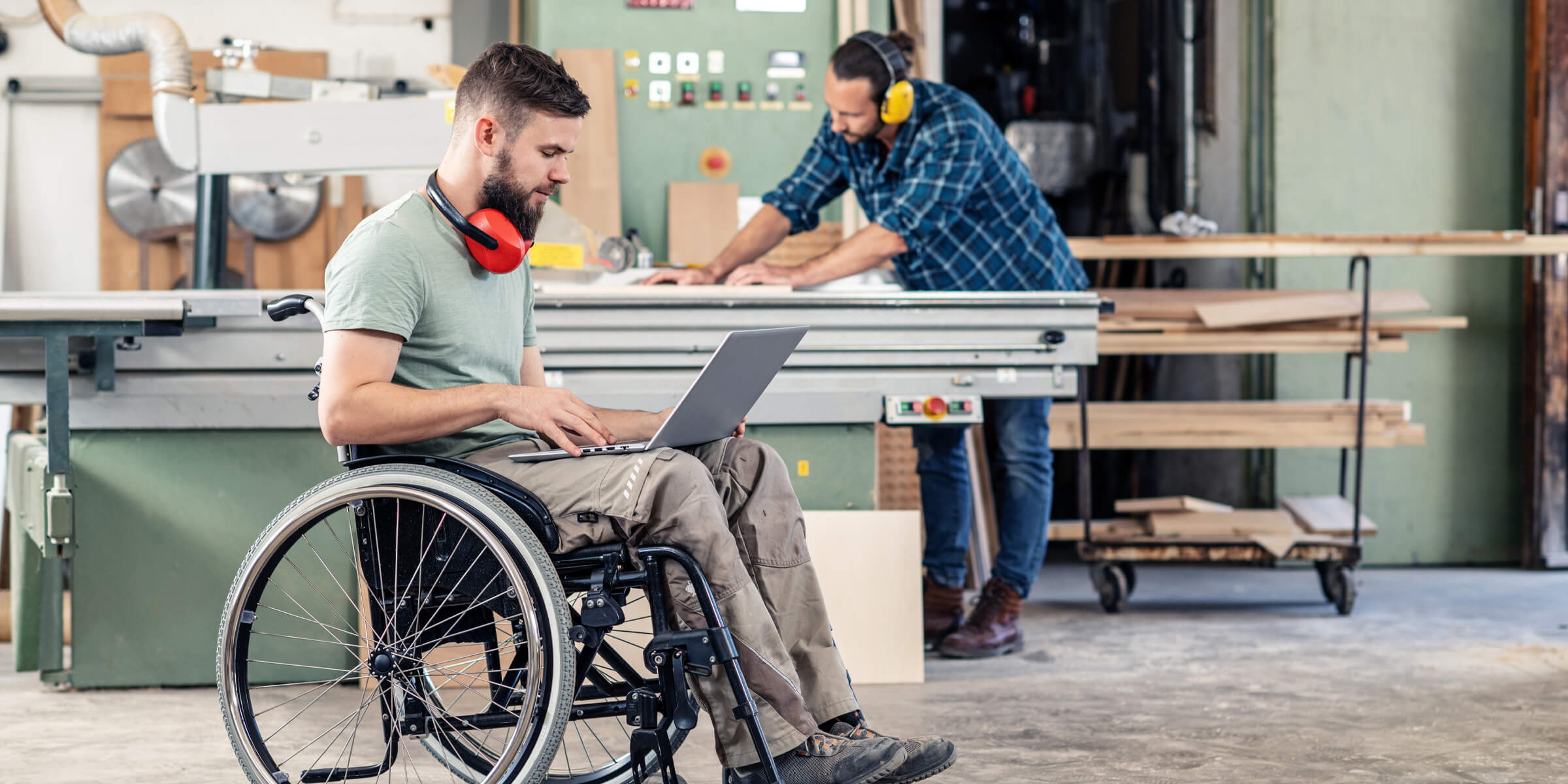 Junger Mensch sitzt im Rollstuhl und schaut in seinen Laptop, im Hintergrund sieht man, dass er in einer Werkstatt ist und ein anderer junger Mensch dort an einer Werkbank mit Holz arbeitet